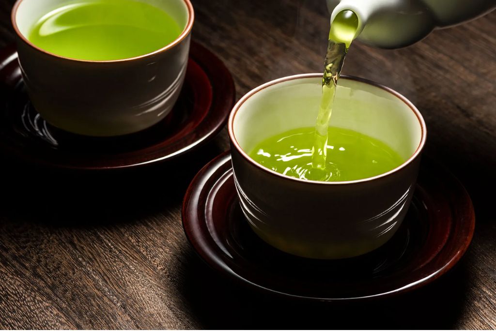 Two cups of green tea on dark wooden surface, with tea being poured into one cup.
