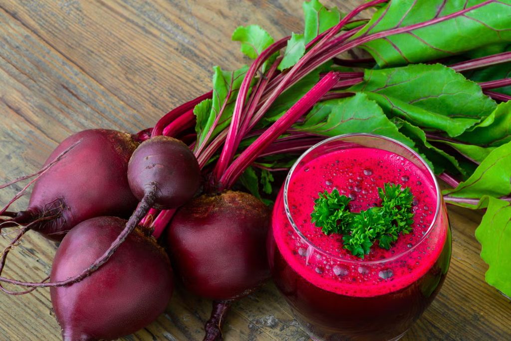 A glass of vibrant pink beet juice garnished with parsley, alongside fresh beets and beet greens on a wooden surface.