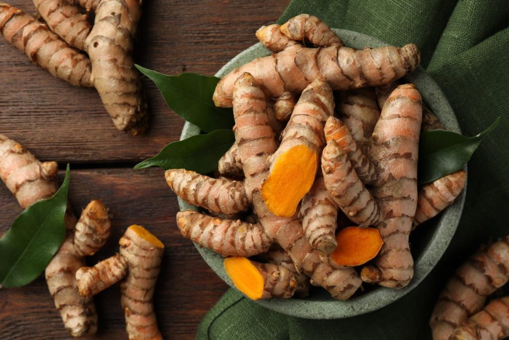 A bowl filled with fresh turmeric roots, some cut to reveal their vibrant orange interior, surrounded by green leaves, on a wooden surface.