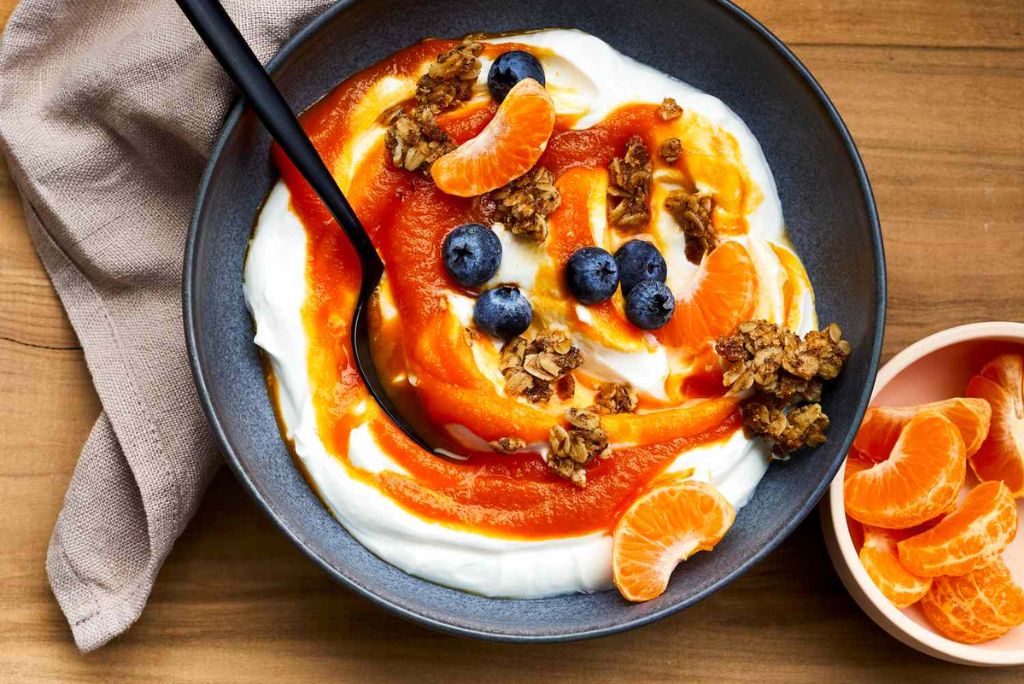 A bowl of yogurt topped with orange segments, blueberries, granola, and a drizzle of orange sauce, accompanied by a small dish of additional orange slices.