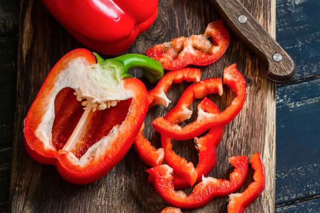 A halved red bell pepper with a green stem sits on a wooden cutting board, alongside slices of red bell pepper and a wooden knife.