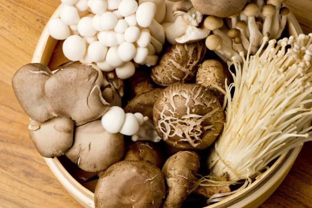 A variety of mushrooms including shiitake, enoki, and white mushrooms arranged in a bamboo basket on a wooden surface.