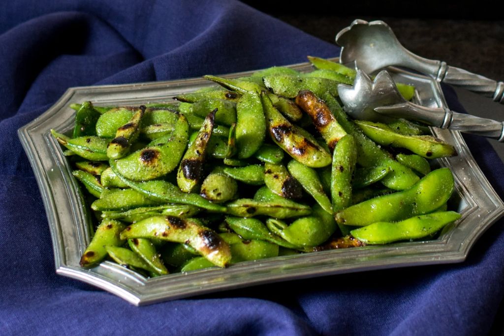 A silver platter filled with grilled edamame beans, garnished with a sprinkle of seasoning, set against a dark blue tablecloth.