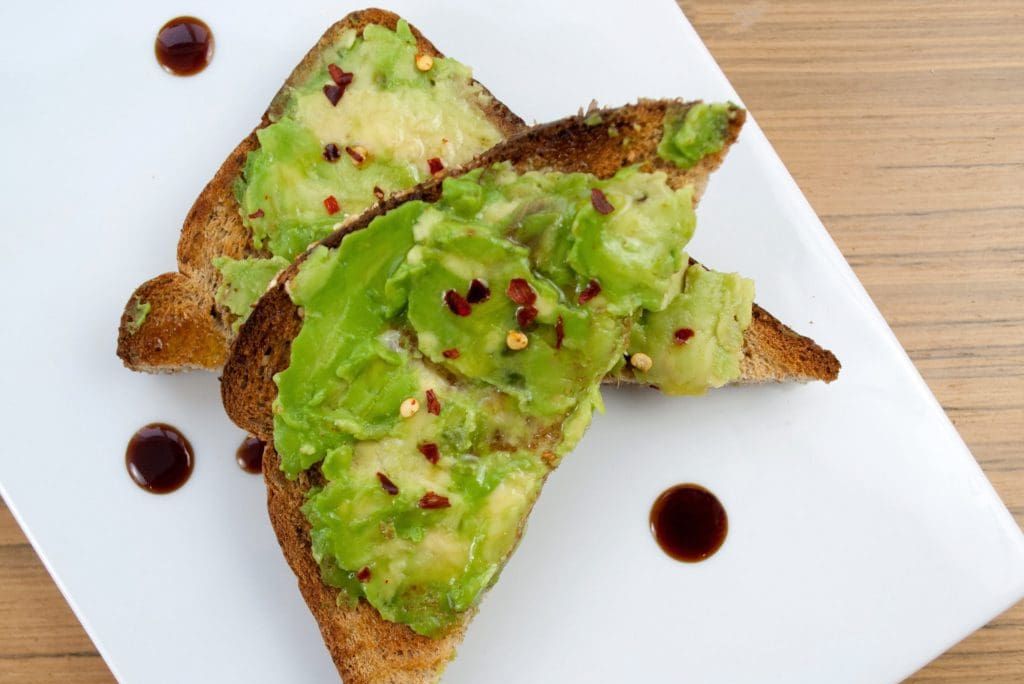 Sliced toasted bread topped with mashed avocado and red pepper flakes, served on a white plate with a drizzle of balsamic reduction.