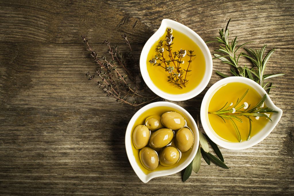 Three small white dishes containing olive oil, herbs, and green olives on a rustic wooden surface.